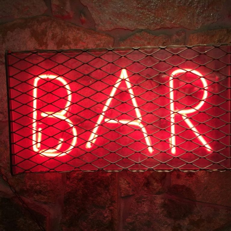 Bar Sign Neon sign reading "BAR" enclosed in a metal mesh frame against a stone wall.
